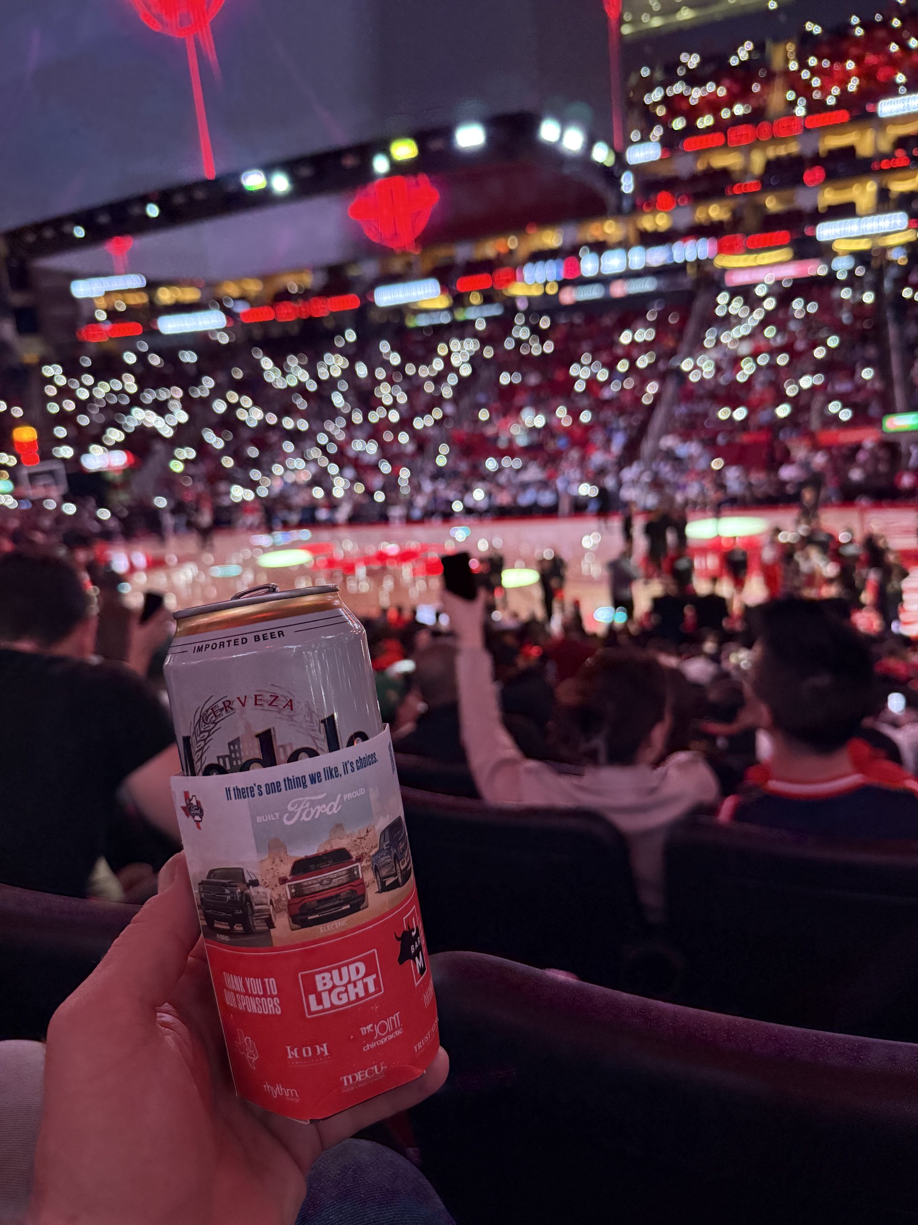 Fan holding a multi-sponsor branded koozie inside a packed live arena at night