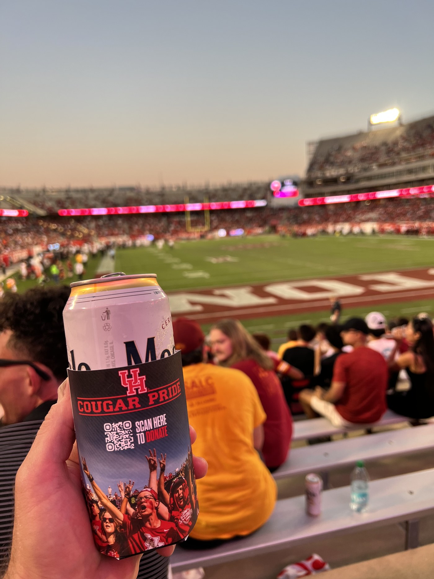 Fan holding a branded koozie with a scan-to-donate QR code inside a college football stadium